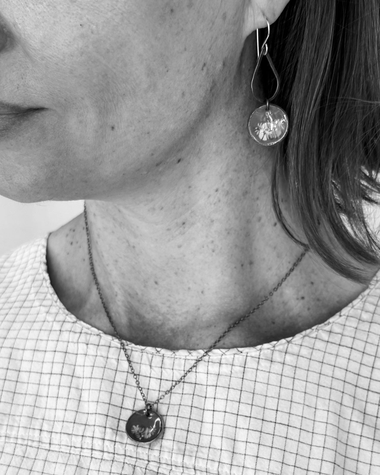 A close-up black and white image of a person's neck and chest area, featuring a necklace with a pendant and a pair of dangling earrings.