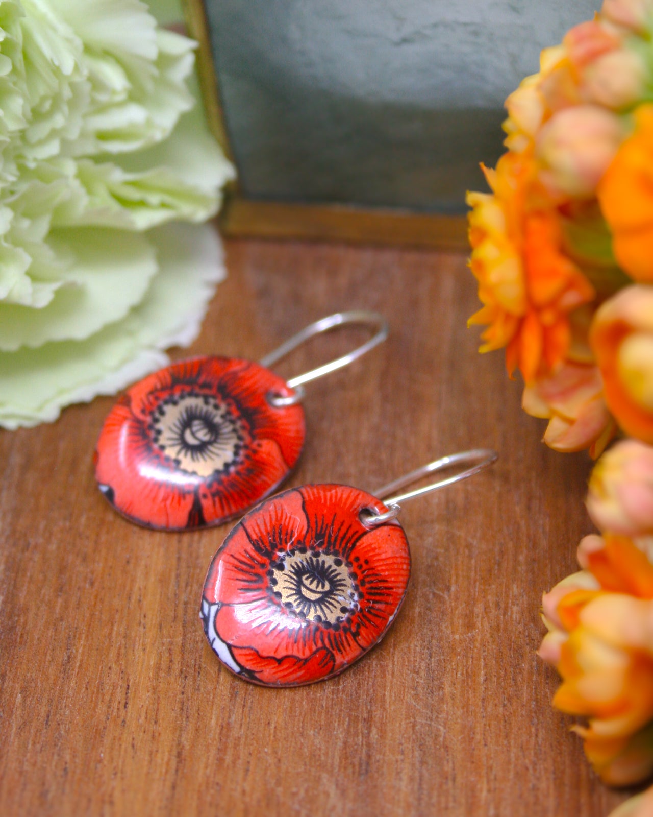 a pair of red and black floral earrings resting on a wooden surface, with a blurred background of orange and white flowers.