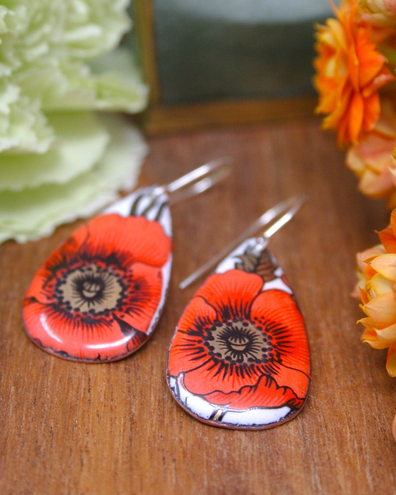 a pair of earrings with a floral design, including red and orange flowers, placed on a wooden surface.