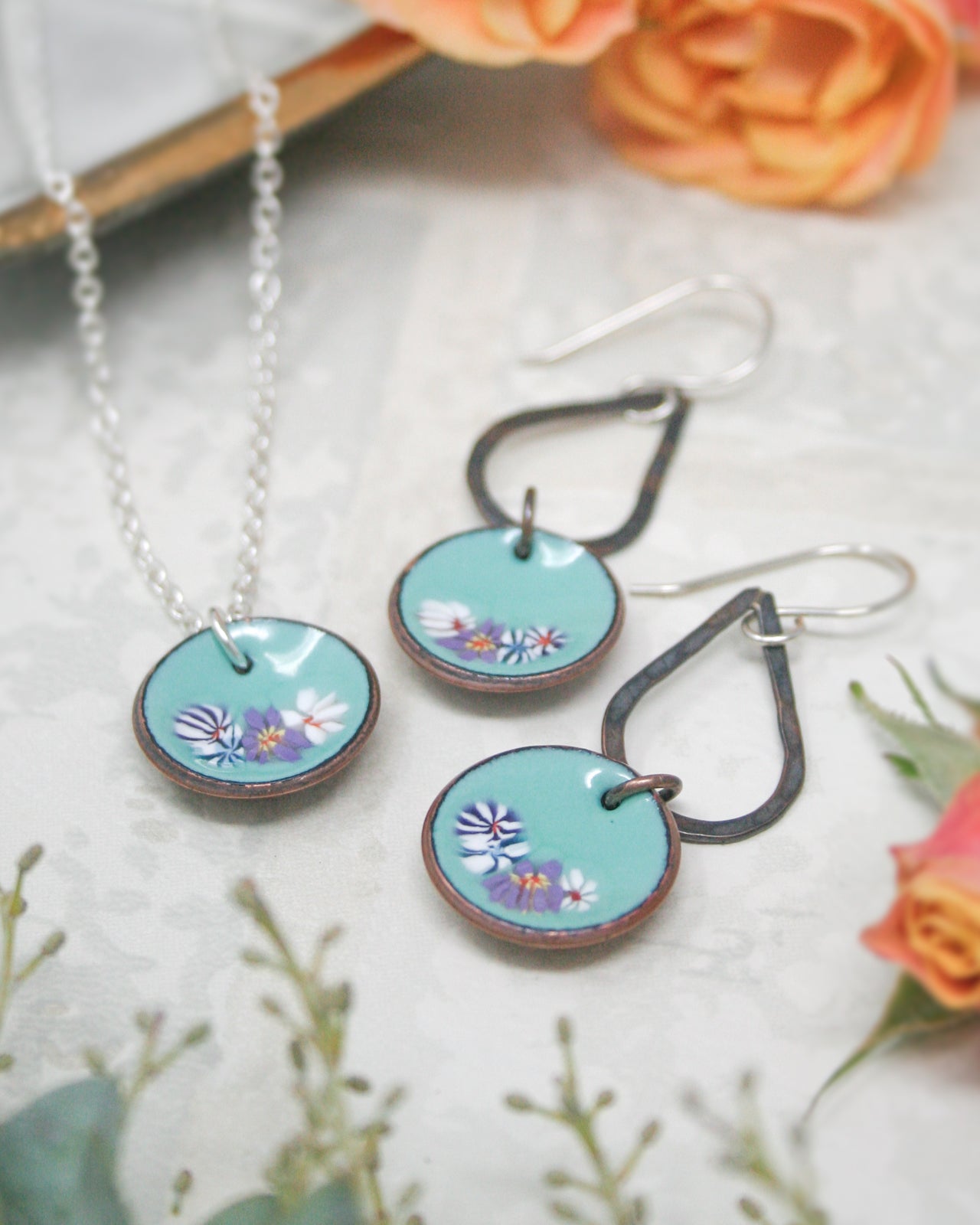 a necklace and earrings with blue and white floral designs, placed on a white surface with orange flowers in the background.