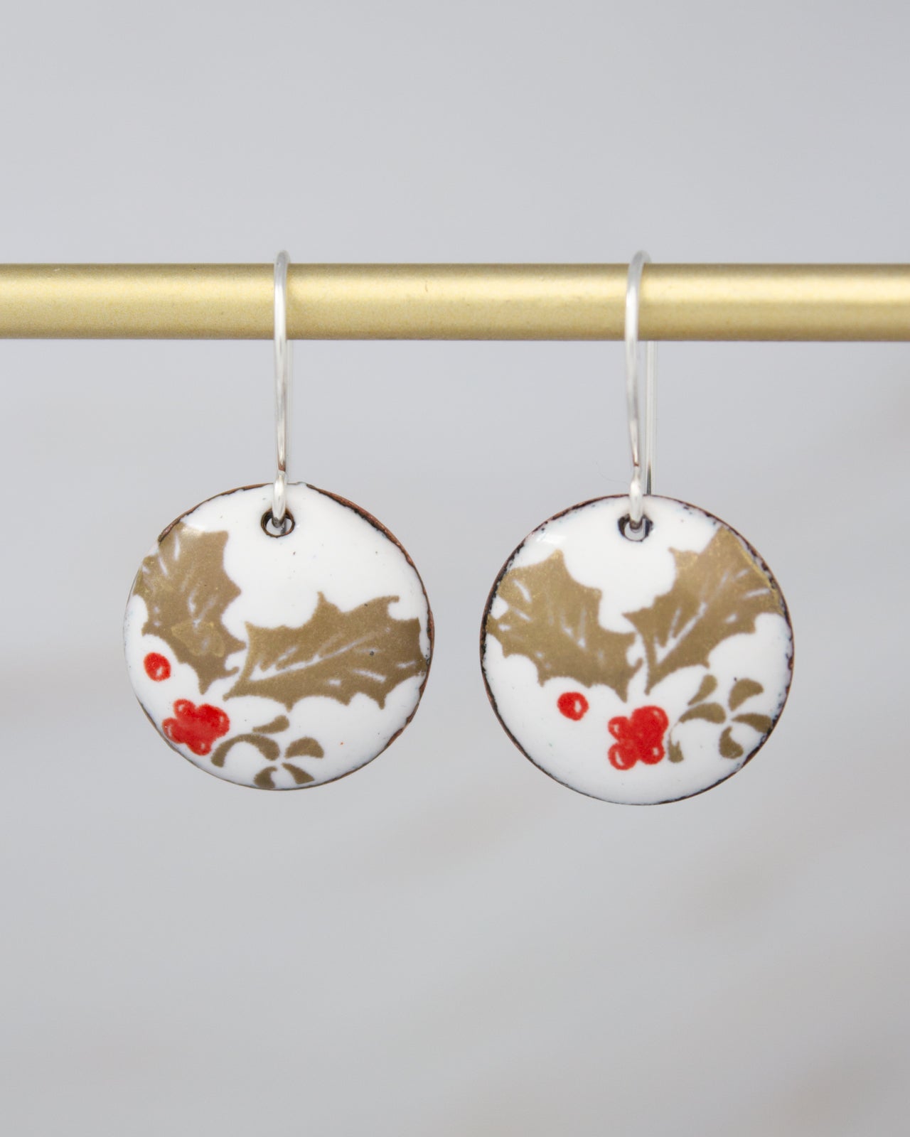 A pair of round earrings with a white and gold floral design, featuring red berries and leaves.