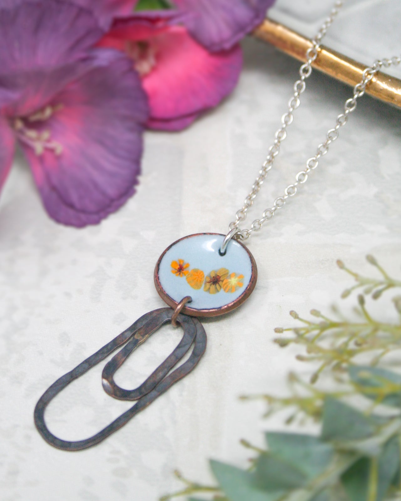 A necklace with a circular pendant featuring a floral design, surrounded by a chain and a purple flower in the background.