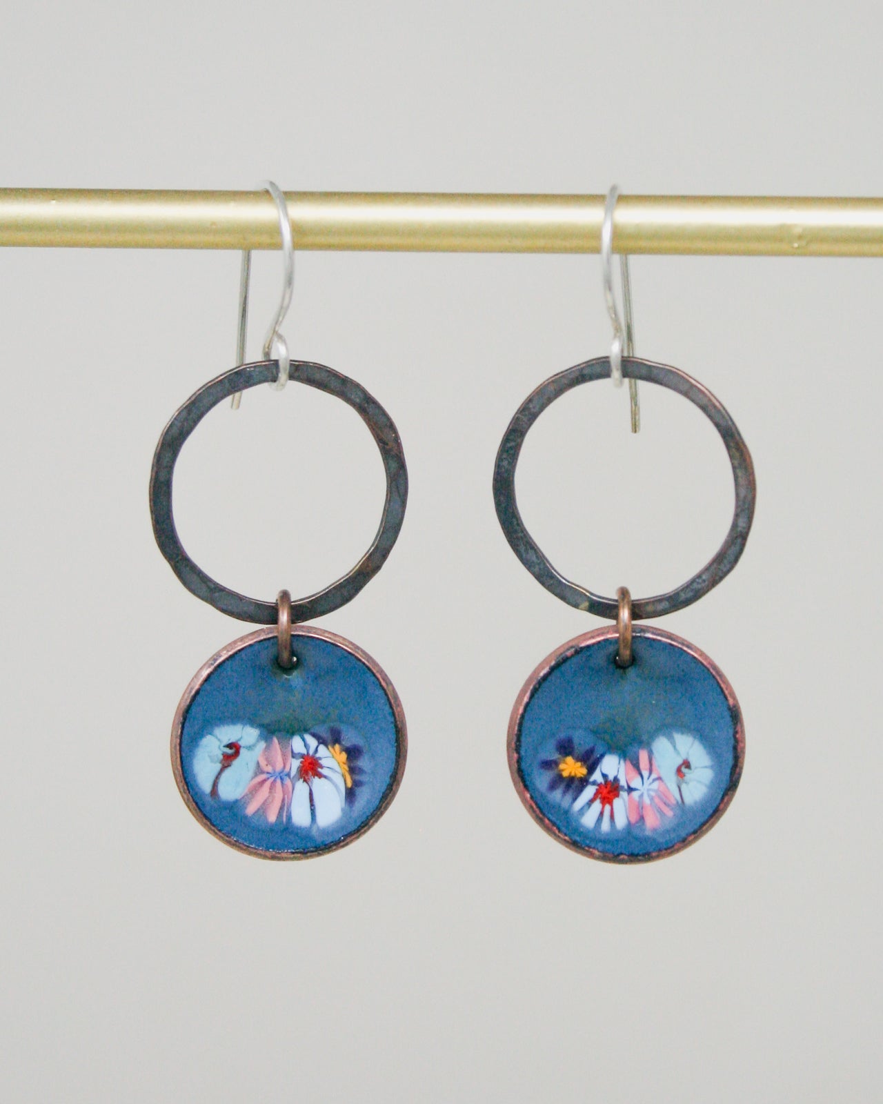 A pair of earrings with blue and pink floral designs on circular pendants, hanging from a gold-colored metal hook.