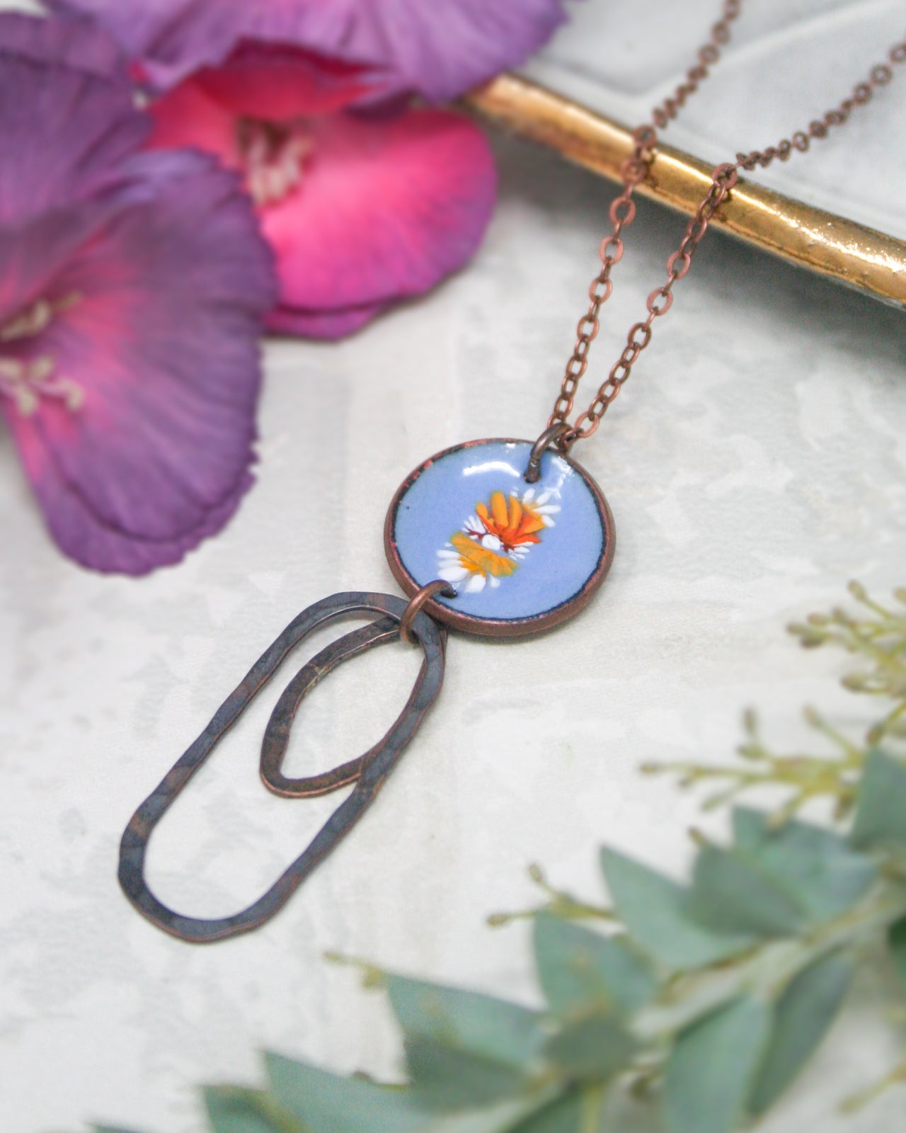 a necklace with a blue circular pendant adorned with orange and white flowers, hanging from a chain. The necklace is set against a backdrop of purple flowers and greenery.