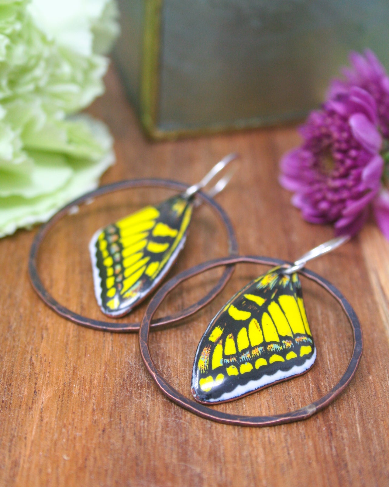 two round earrings with butterfly wing designs, placed on a wooden surface alongside a purple flower.