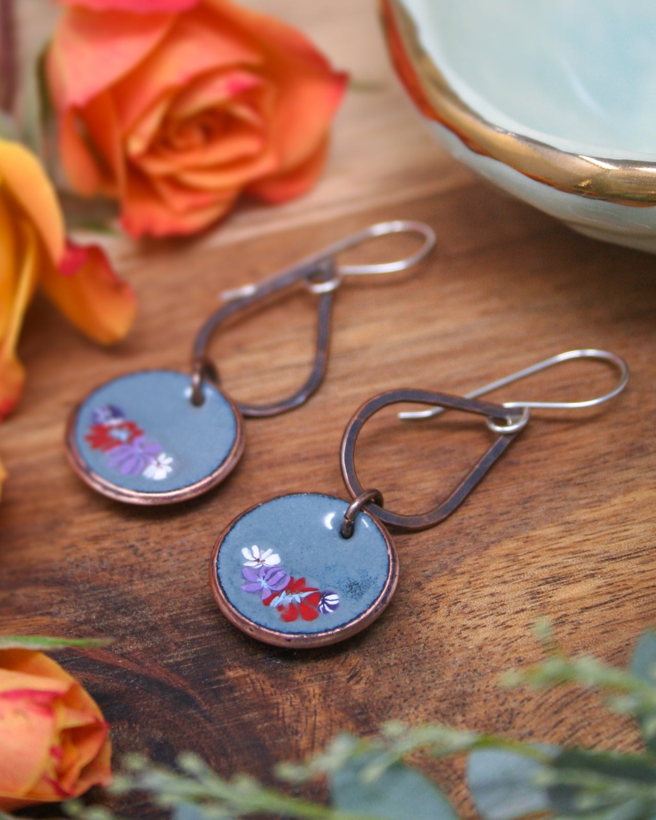 a pair of earrings with a floral design, placed on a wooden surface alongside a bowl and some orange flowers.