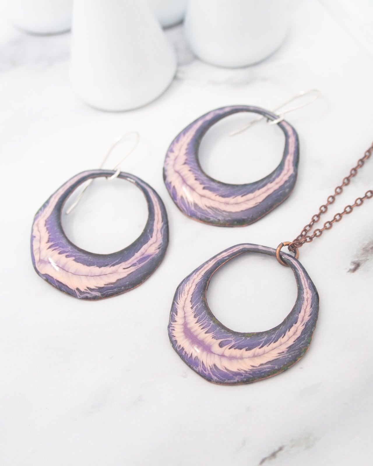 a pair of purple and pink oval-shaped earrings and a necklace with a chain, all displayed on a white marble surface.