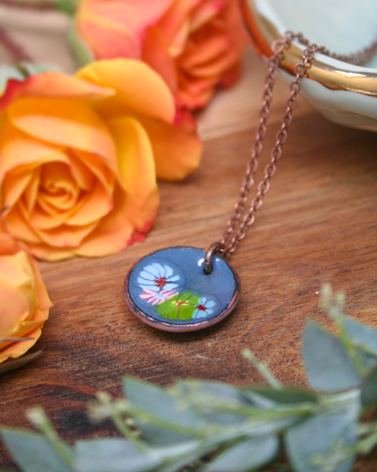 a necklace with a blue pendant adorned with flowers, resting on a wooden surface. The necklace is surrounded by vibrant orange and yellow roses, creating a visually appealing and colorful composition.