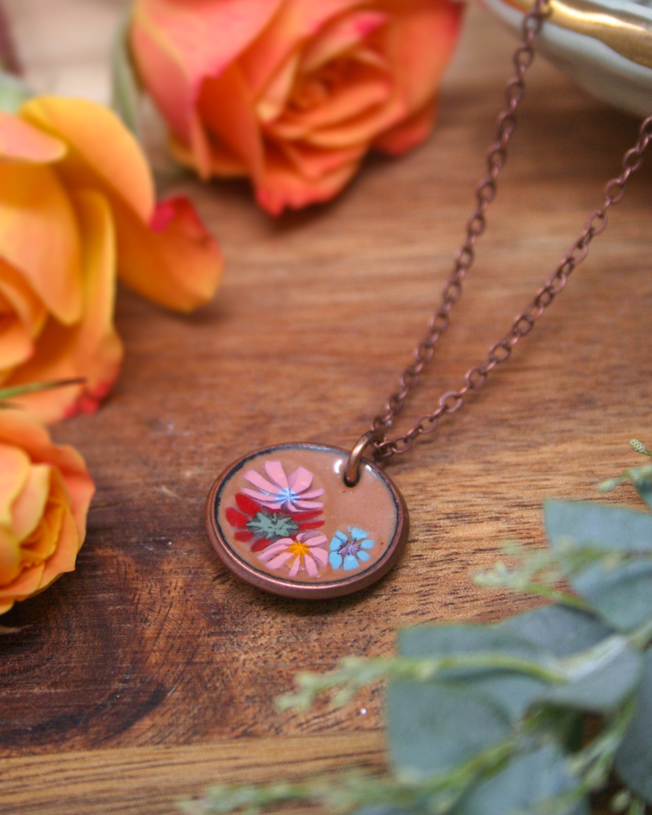 A copper-colored pendant necklace with a circular design featuring a floral pattern rests on a wooden surface, surrounded by vibrant orange and yellow roses.