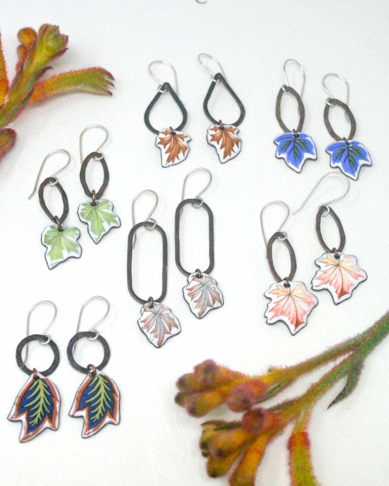 a collection of earrings with leaf-shaped designs, arranged on a white background alongside some plant parts.