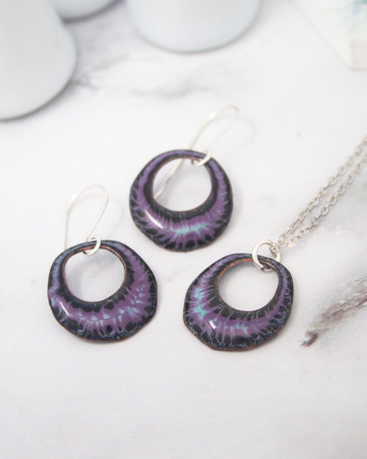 A pair of purple earrings and a necklace with a pendant are displayed on a white surface.
