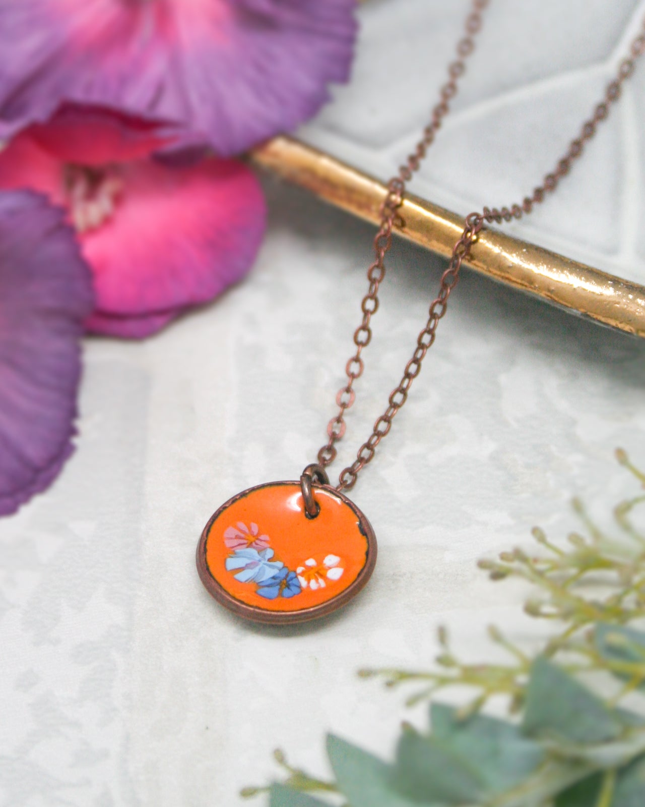 a necklace with a circular pendant, which is an orange-colored piece of jewelry. The pendant has a floral design on it. The necklace is placed on a white surface, and there are purple flowers in the background.