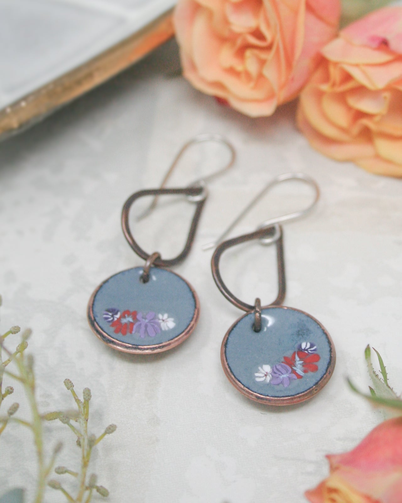 A pair of blue and copper earrings with floral designs, placed on a white surface next to pink roses.