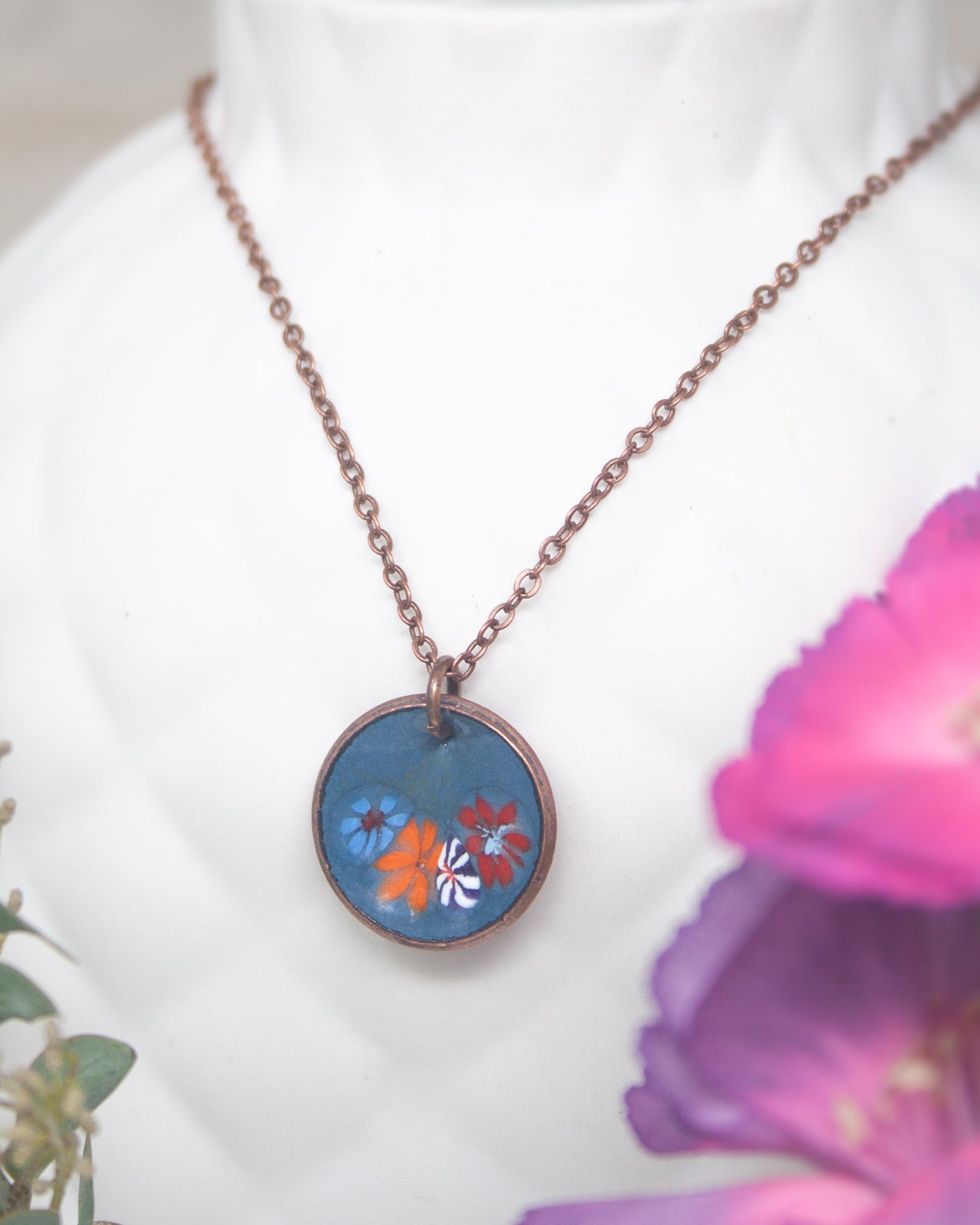 A round pendant necklace with a blue and orange floral design, displayed on a white mannequin or vase.