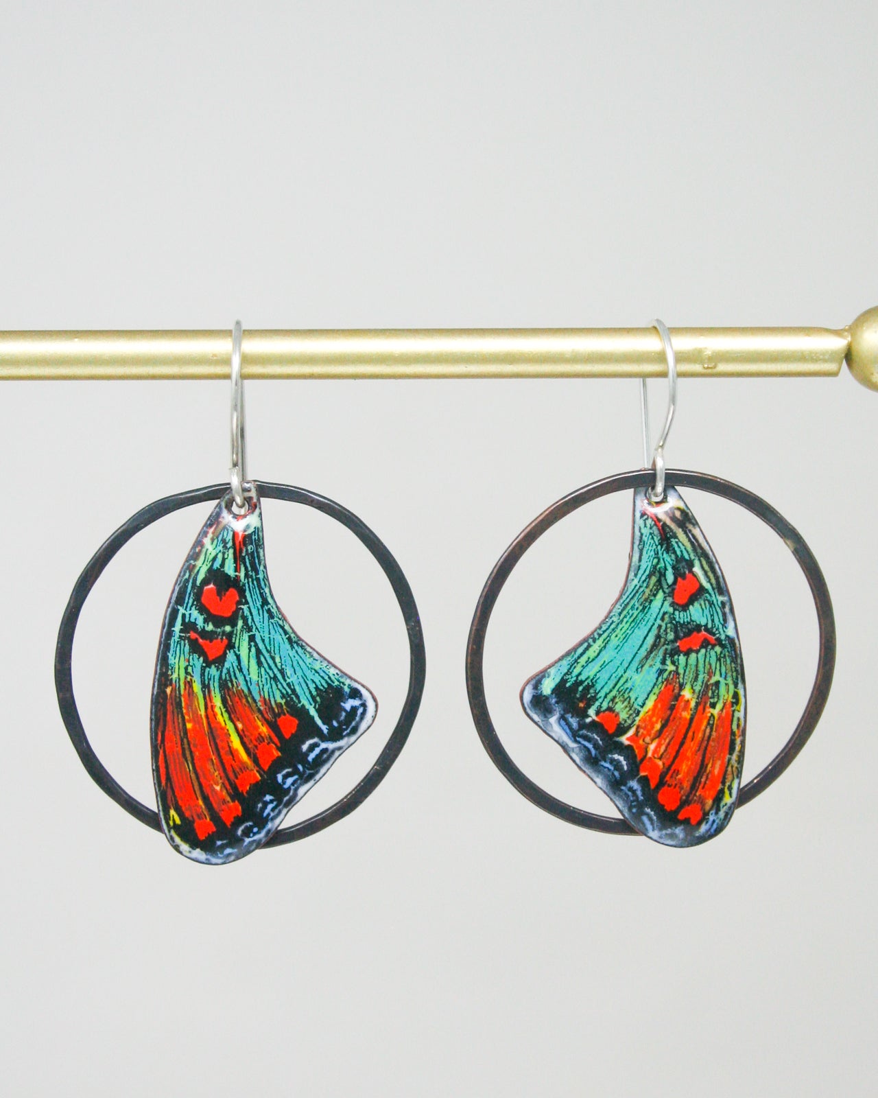 A pair of colorful earrings with butterfly wing designs, hanging from a gold-colored metal hook.