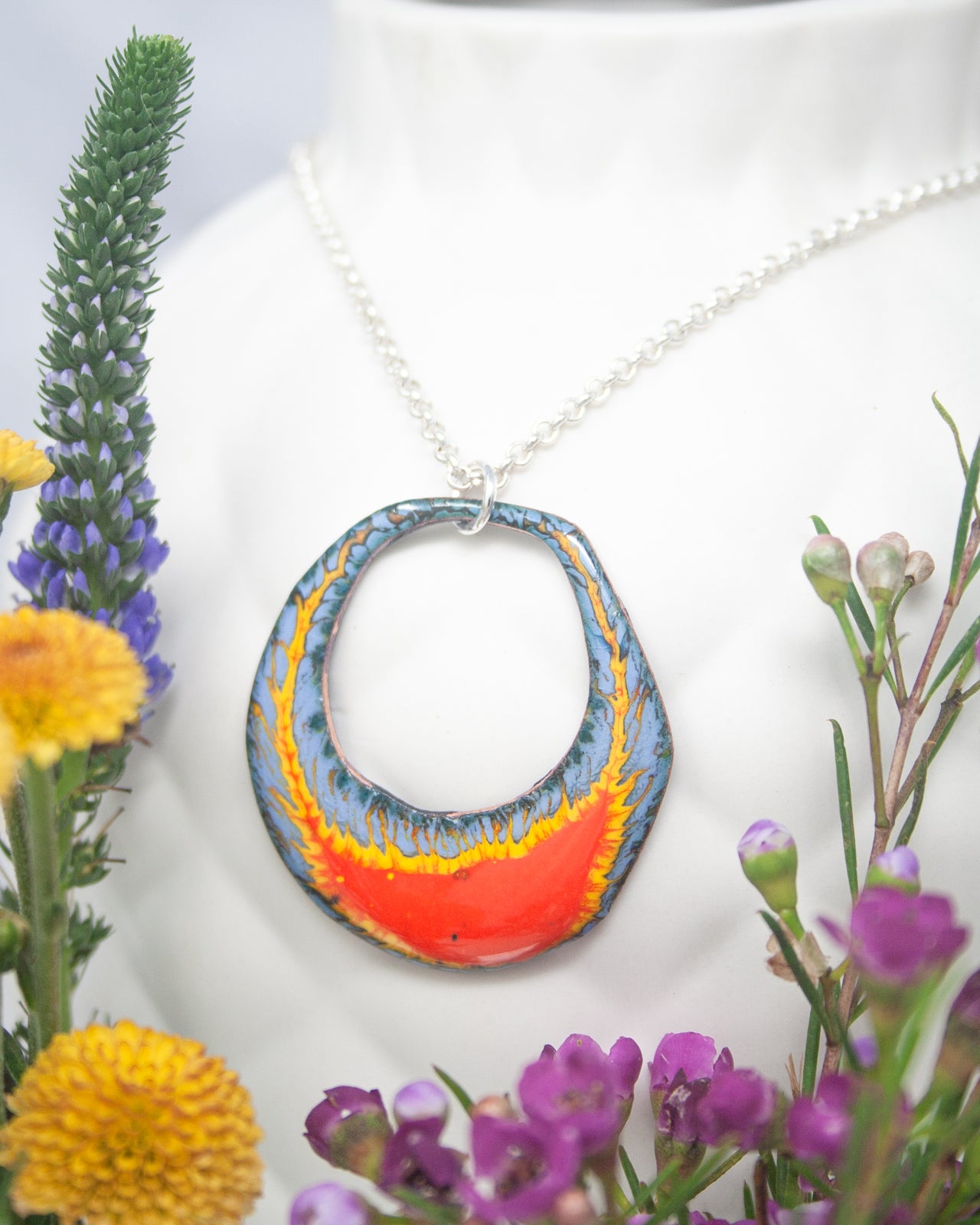 A colorful pendant necklace with a large, circular design hangs from a white bust form, surrounded by vibrant flowers and foliage.