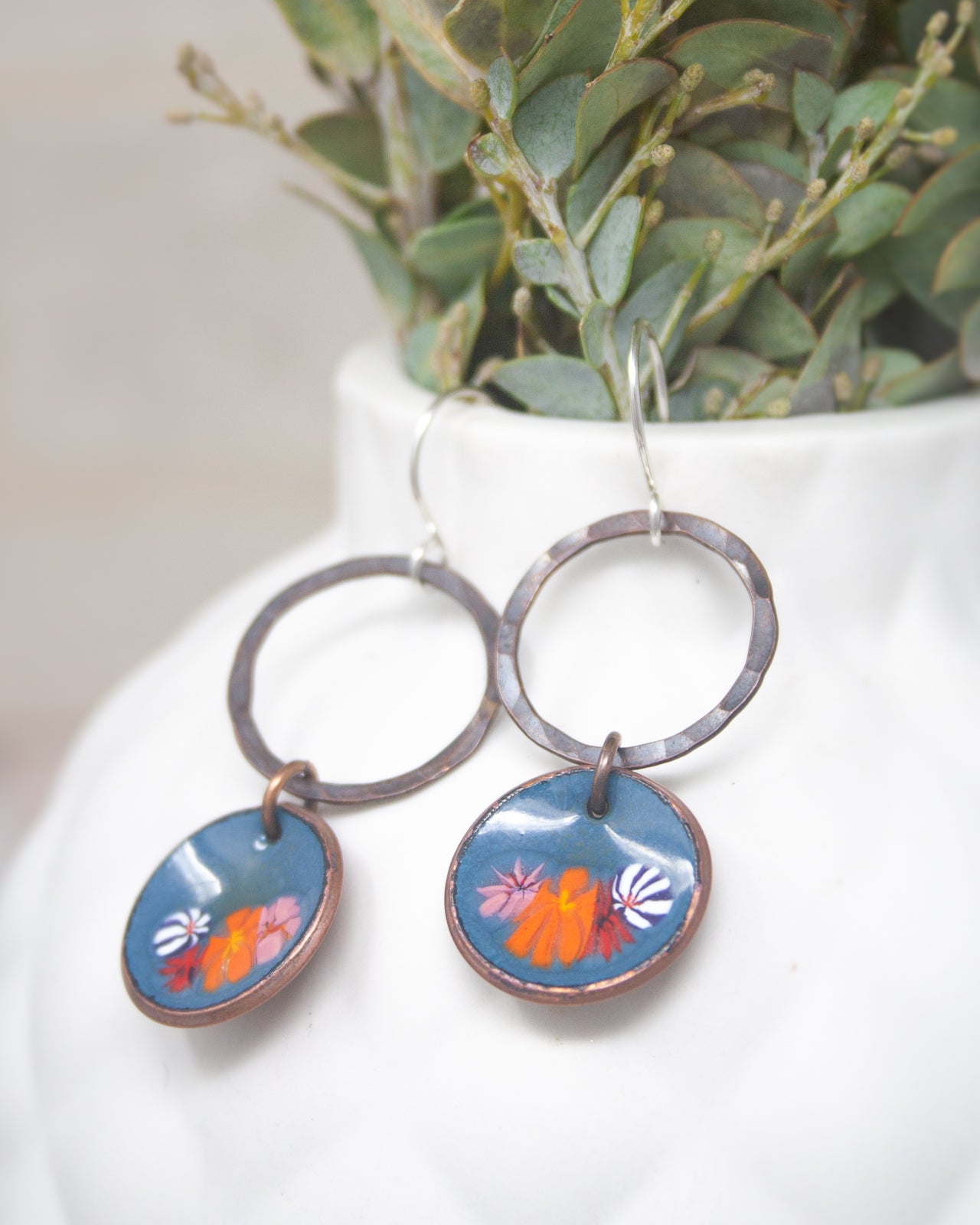 A pair of earrings with blue and orange floral designs, hanging from a white vase with greenery.