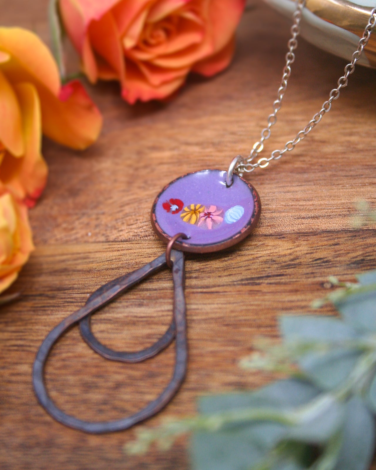 A purple pendant with a floral design hangs from a silver chain, surrounded by orange and yellow flowers on a wooden surface.