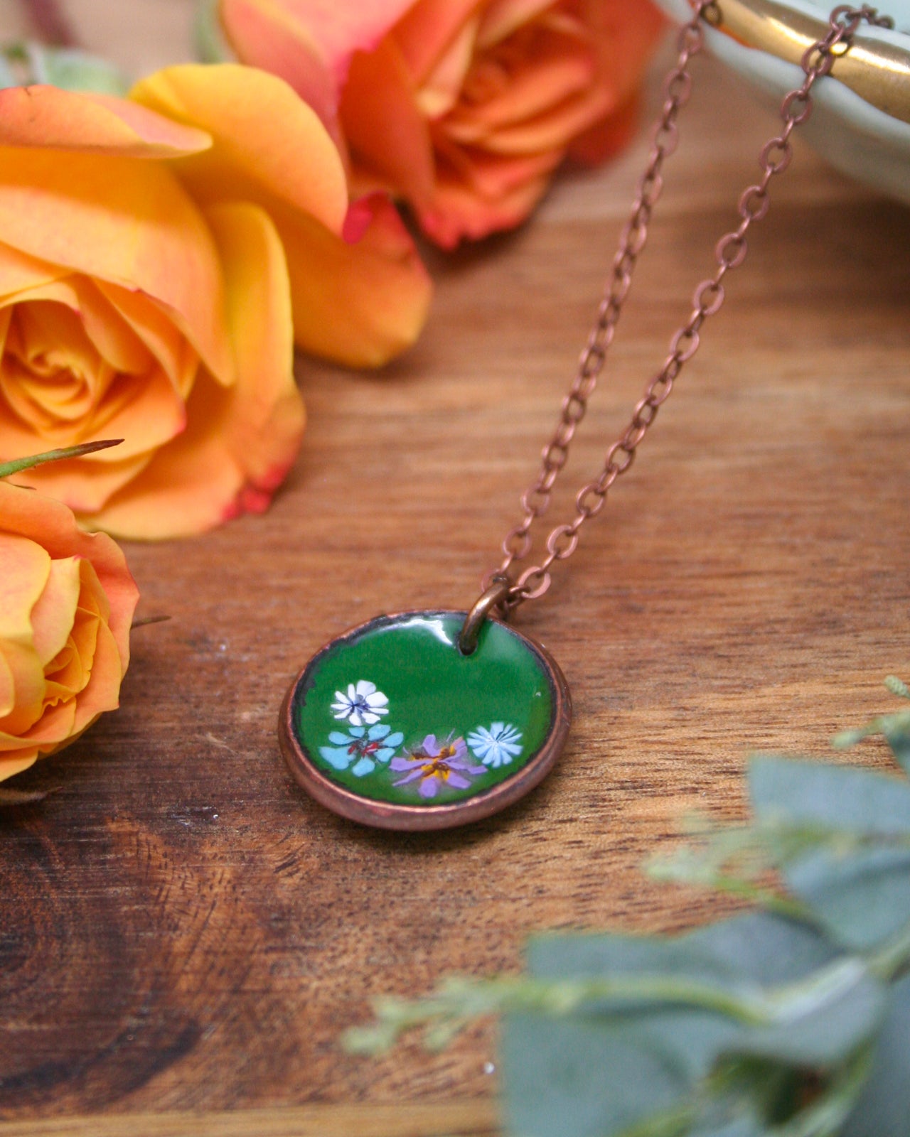 A green pendant necklace with a floral design rests on a wooden surface, surrounded by vibrant orange and yellow roses.