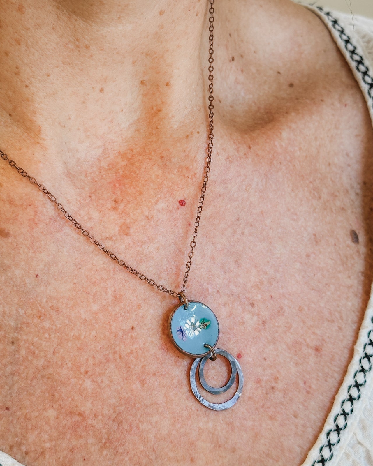 A woman wearing a necklace with a blue circular pendant featuring a floral design.
