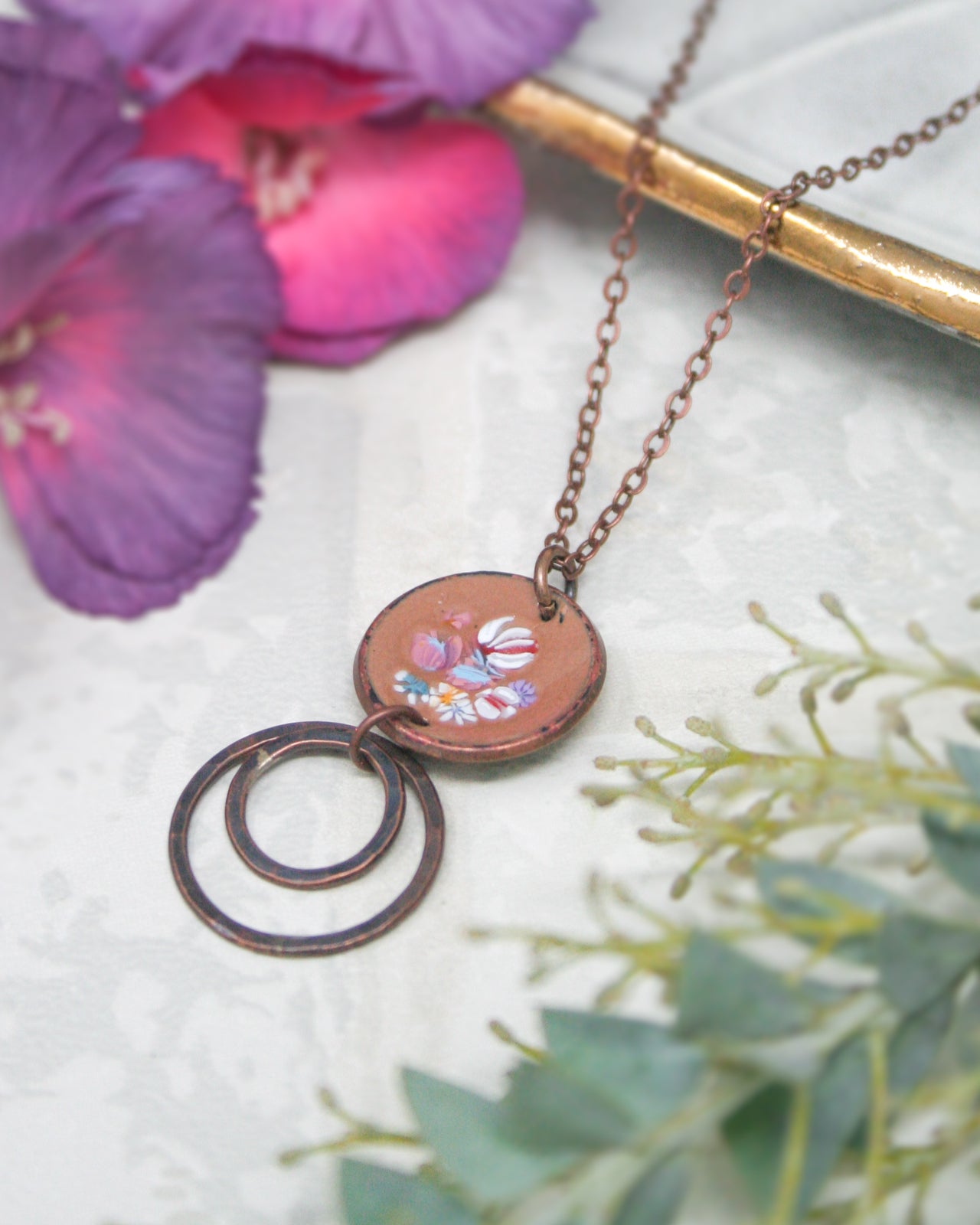 a necklace with a circular pendant, adorned with a floral design, resting on a white surface. The necklace is accompanied by a pair of purple flowers and some greenery, creating a visually appealing and harmonious composition.