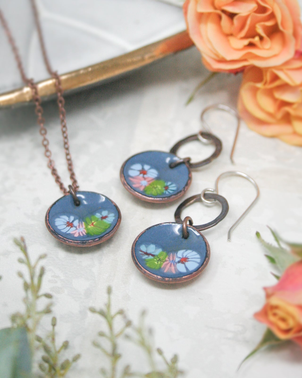 a necklace with a pendant and earrings with a flower design, placed on a white surface with a rose and other flowers in the background.
