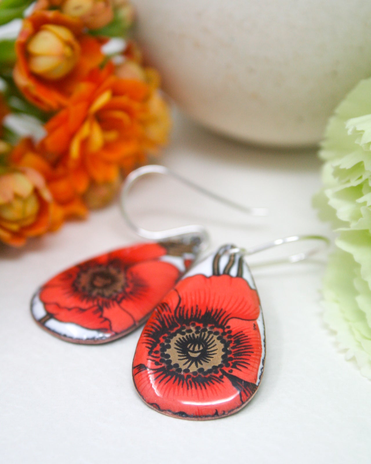 A pair of red and white earrings with a floral design, placed on a white surface next to a bouquet of orange and yellow flowers.