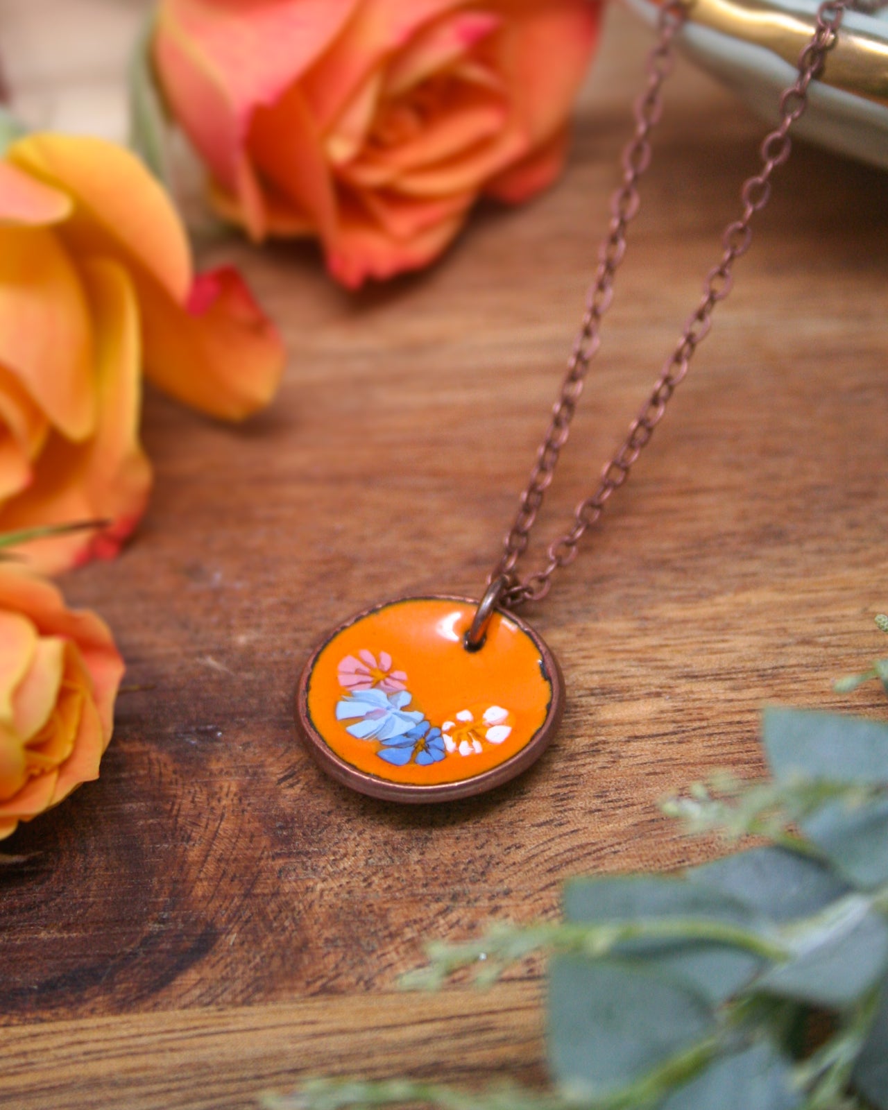 a necklace with an orange pendant adorned with blue and pink flowers, resting on a wooden surface.
