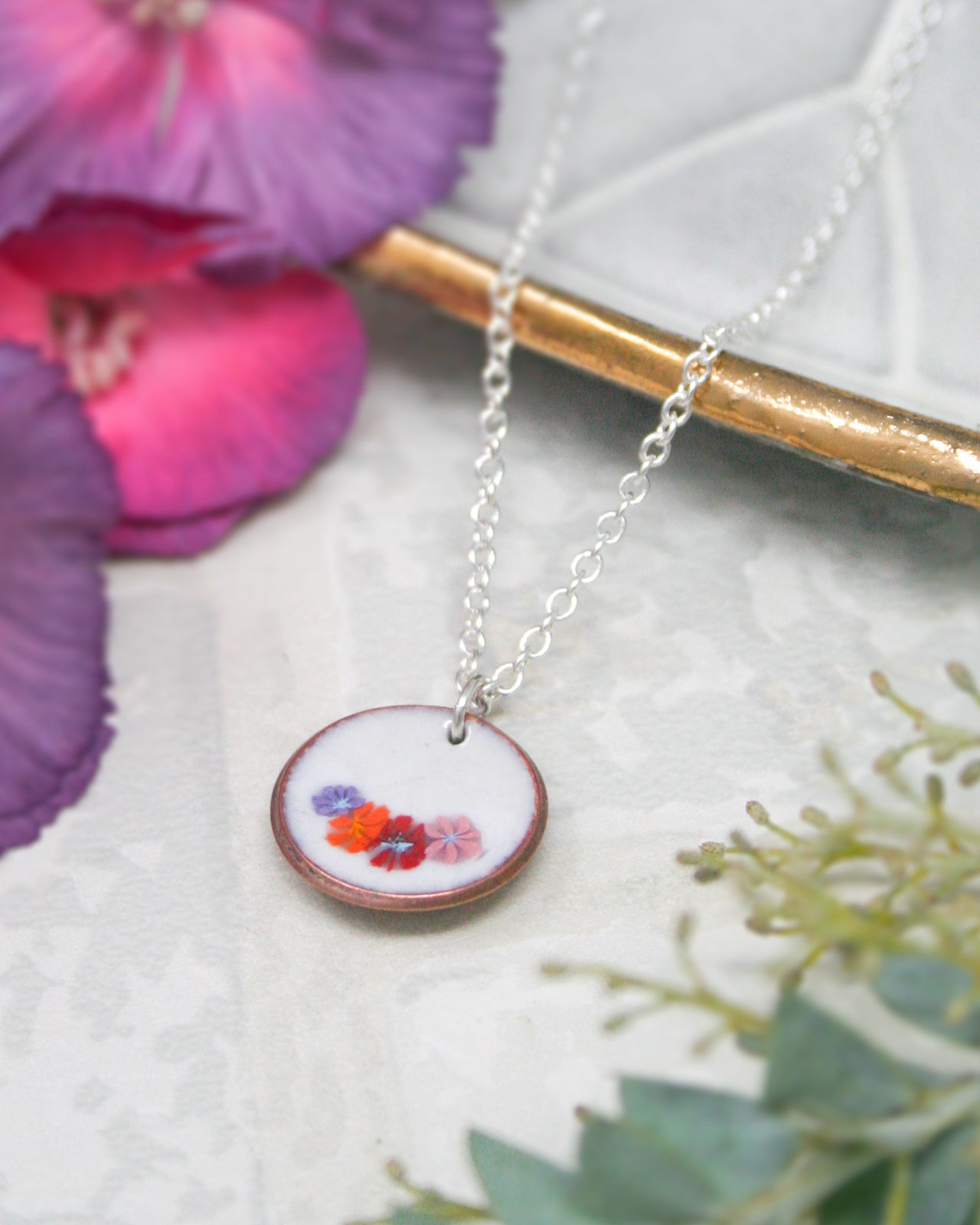A delicate necklace with a round pendant featuring a floral design rests on a white surface, surrounded by vibrant purple and pink flowers.