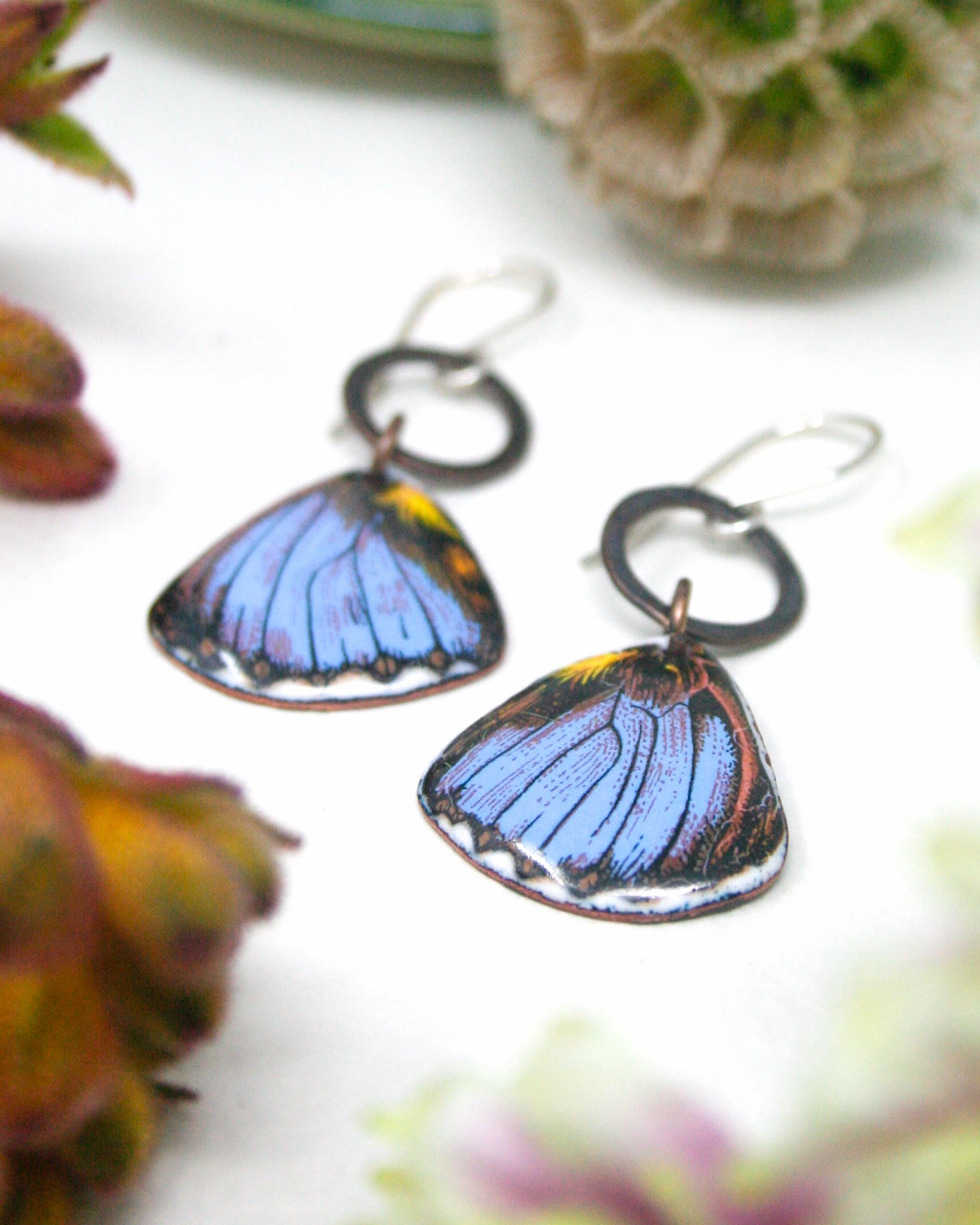 a pair of earrings with butterfly wing designs, placed on a white surface with some blurred flowers in the background.