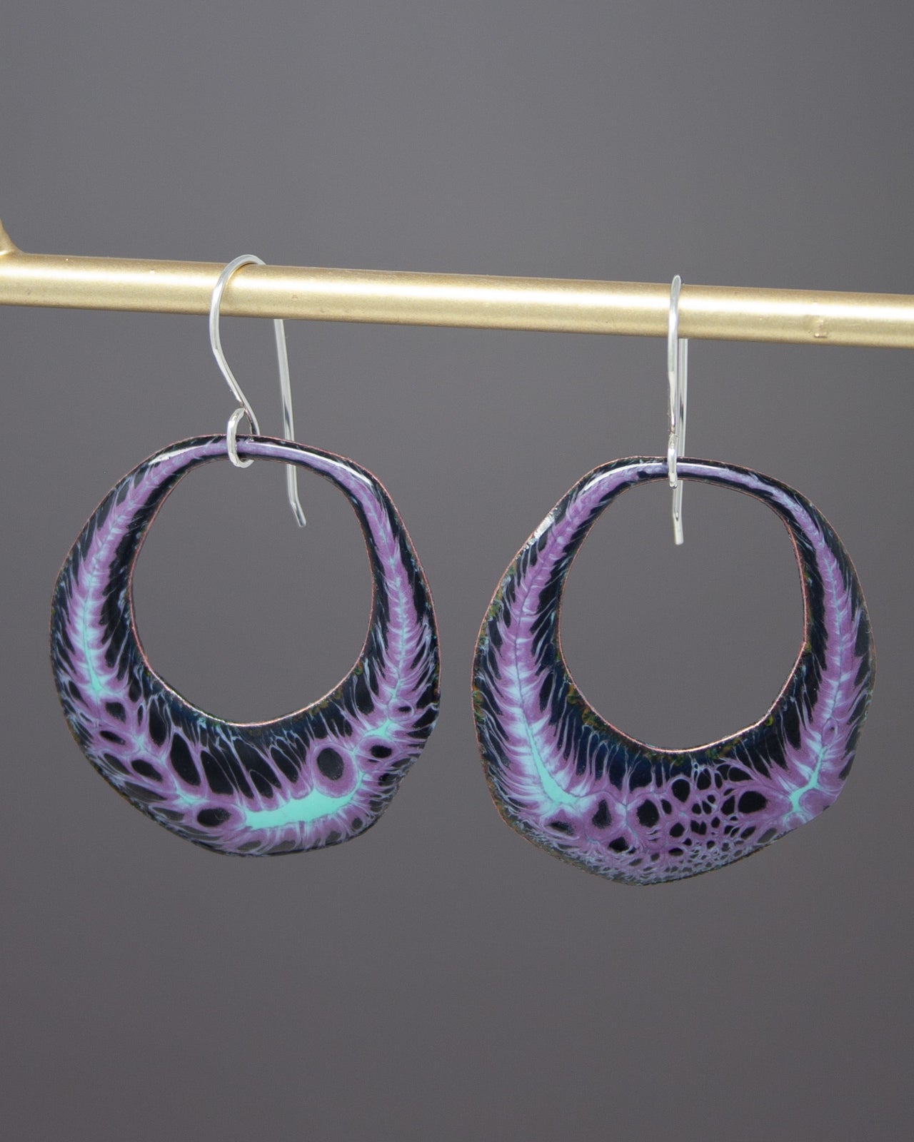 A pair of earrings with a purple and black floral design hanging from a gold-colored metal hook.