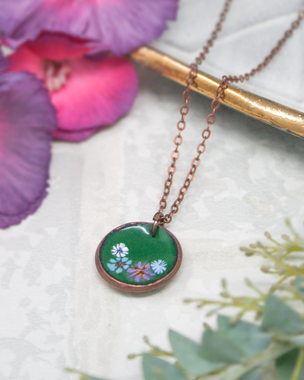A necklace with a green pendant featuring a floral design is displayed on a white surface, accompanied by a purple flower and some greenery.