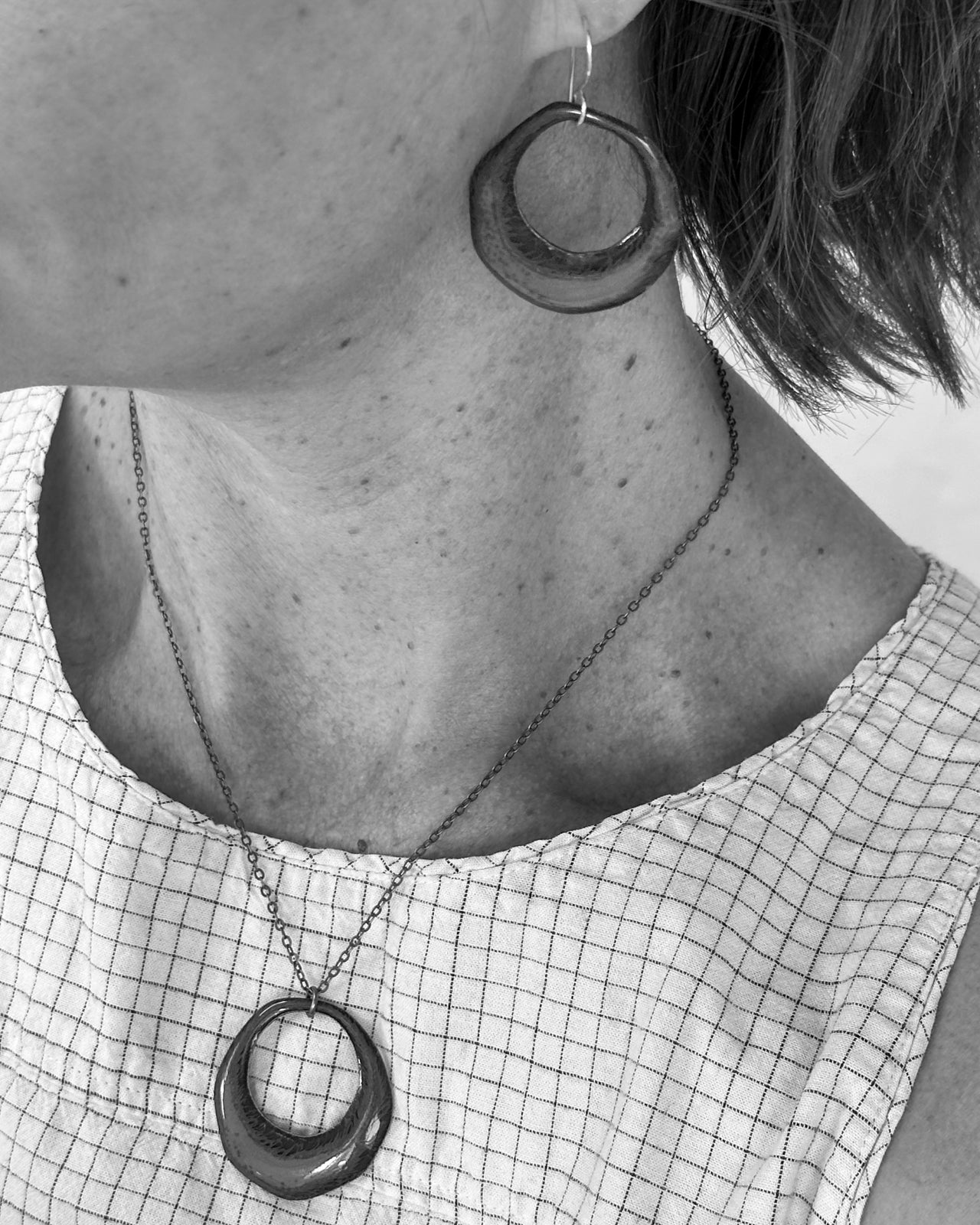 A close-up black and white image of a person's neck and chest area, featuring a necklace with a pendant and a pair of hoop earrings.