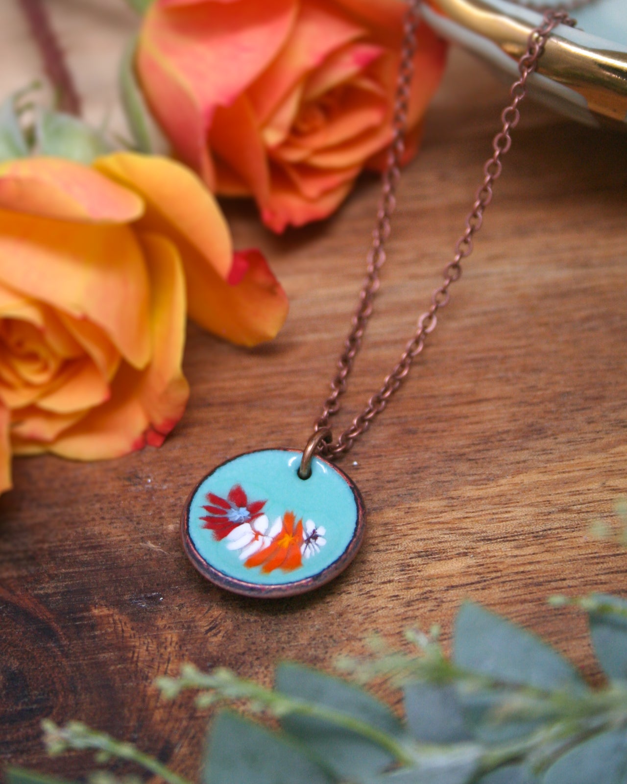 A necklace with a circular pendant featuring a floral design rests on a wooden surface, surrounded by vibrant orange and yellow roses.