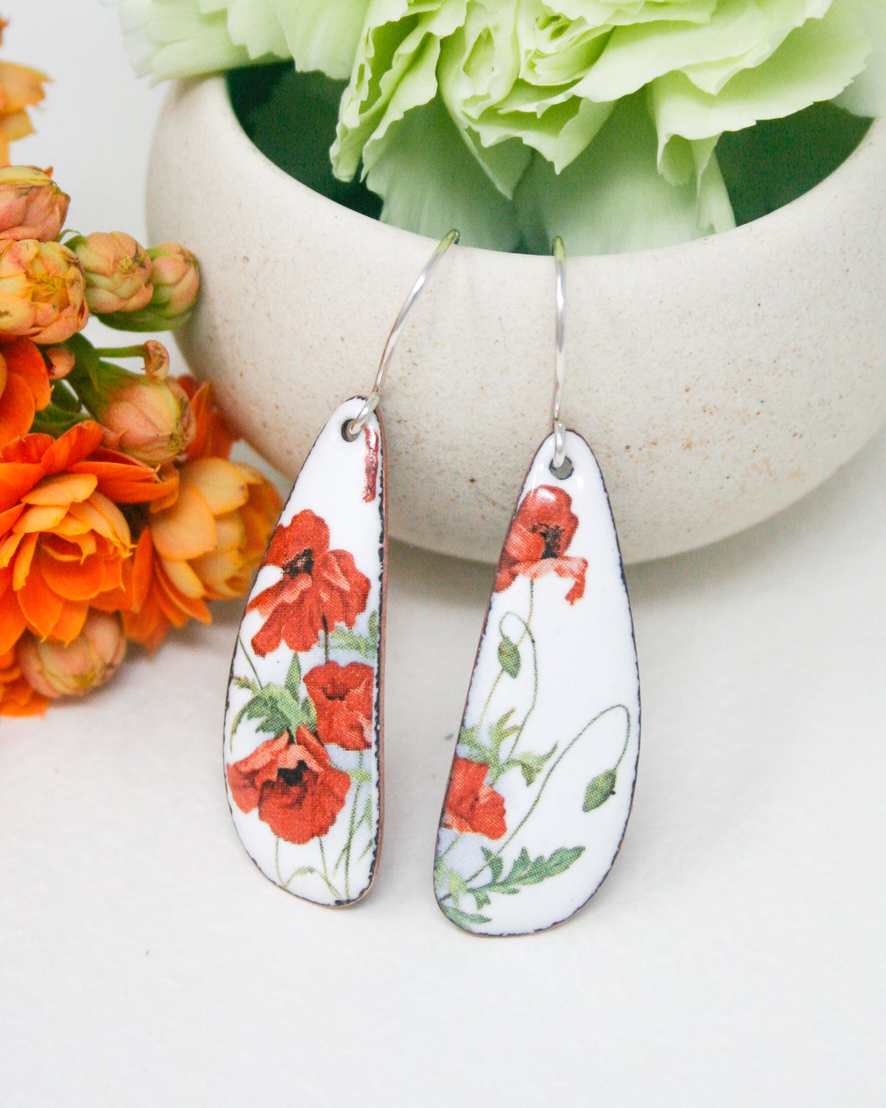A pair of white earrings with red flowers on them, placed on a white surface next to a white vase filled with green flowers.