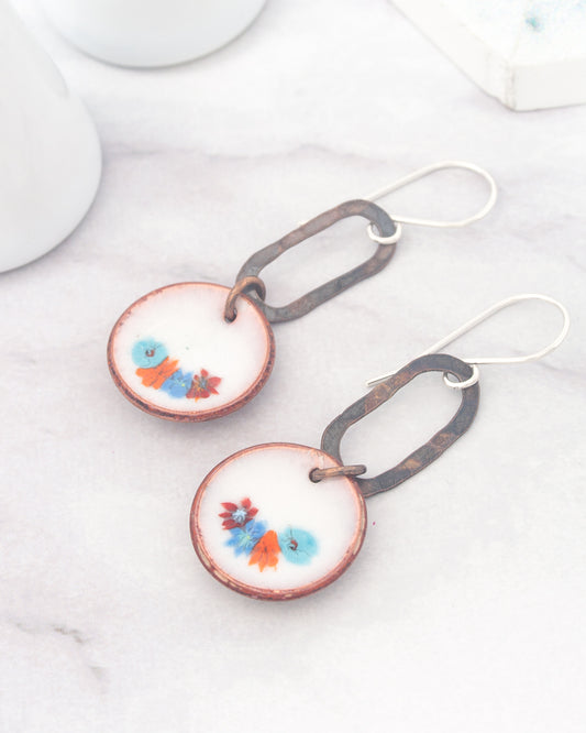 A pair of earrings with a floral design on the circular pendants, resting on a marble surface.