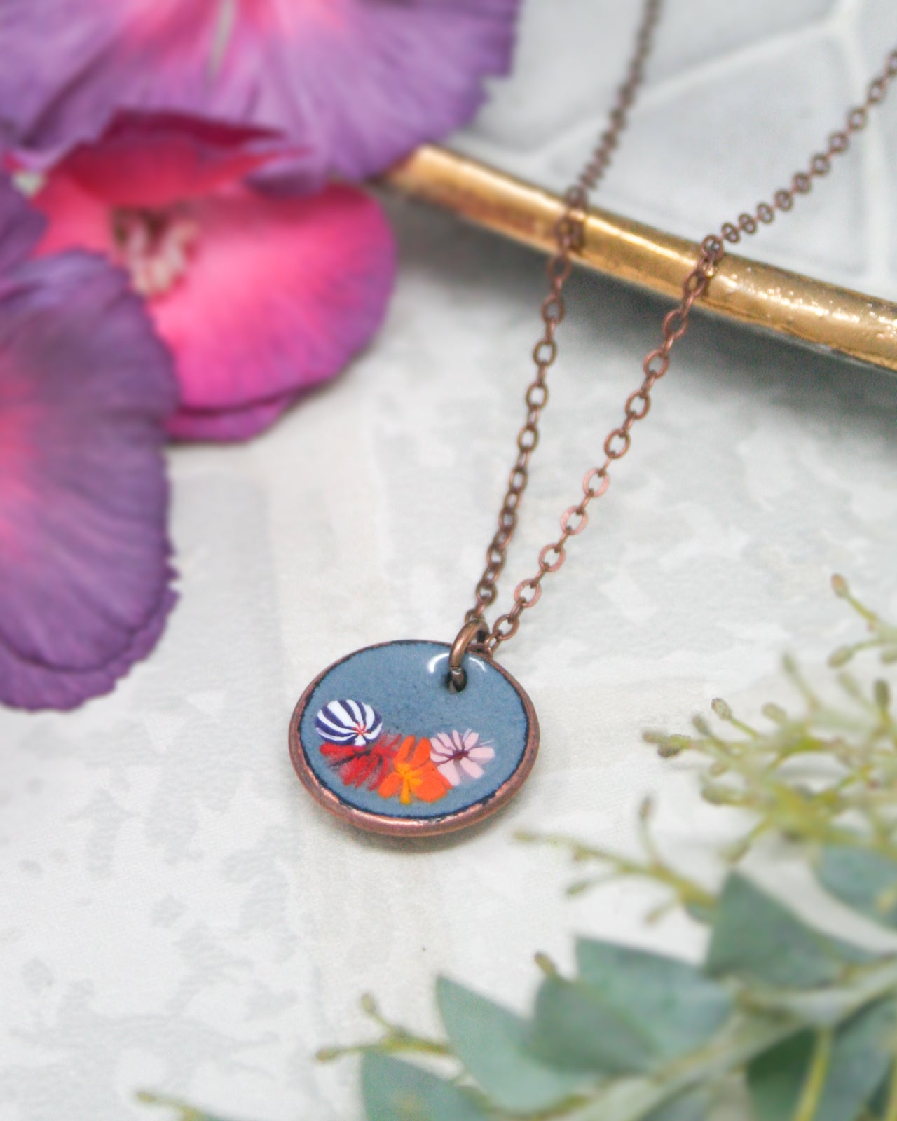 A necklace with a circular pendant featuring a floral design is displayed on a white surface, surrounded by purple and green flowers.