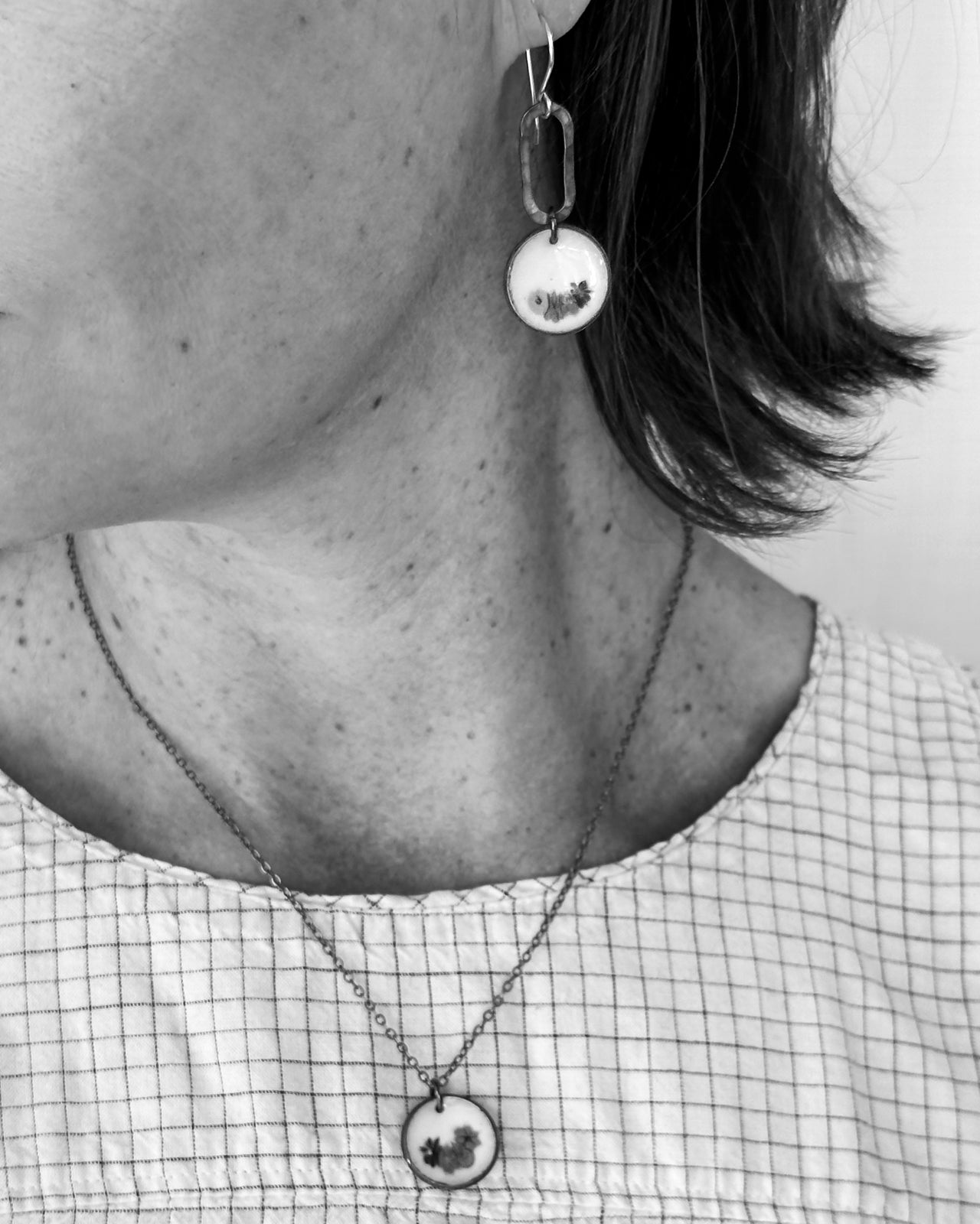 A close-up black and white image of a person's neck and chest area, featuring a necklace with a pendant and a pair of dangling earrings.