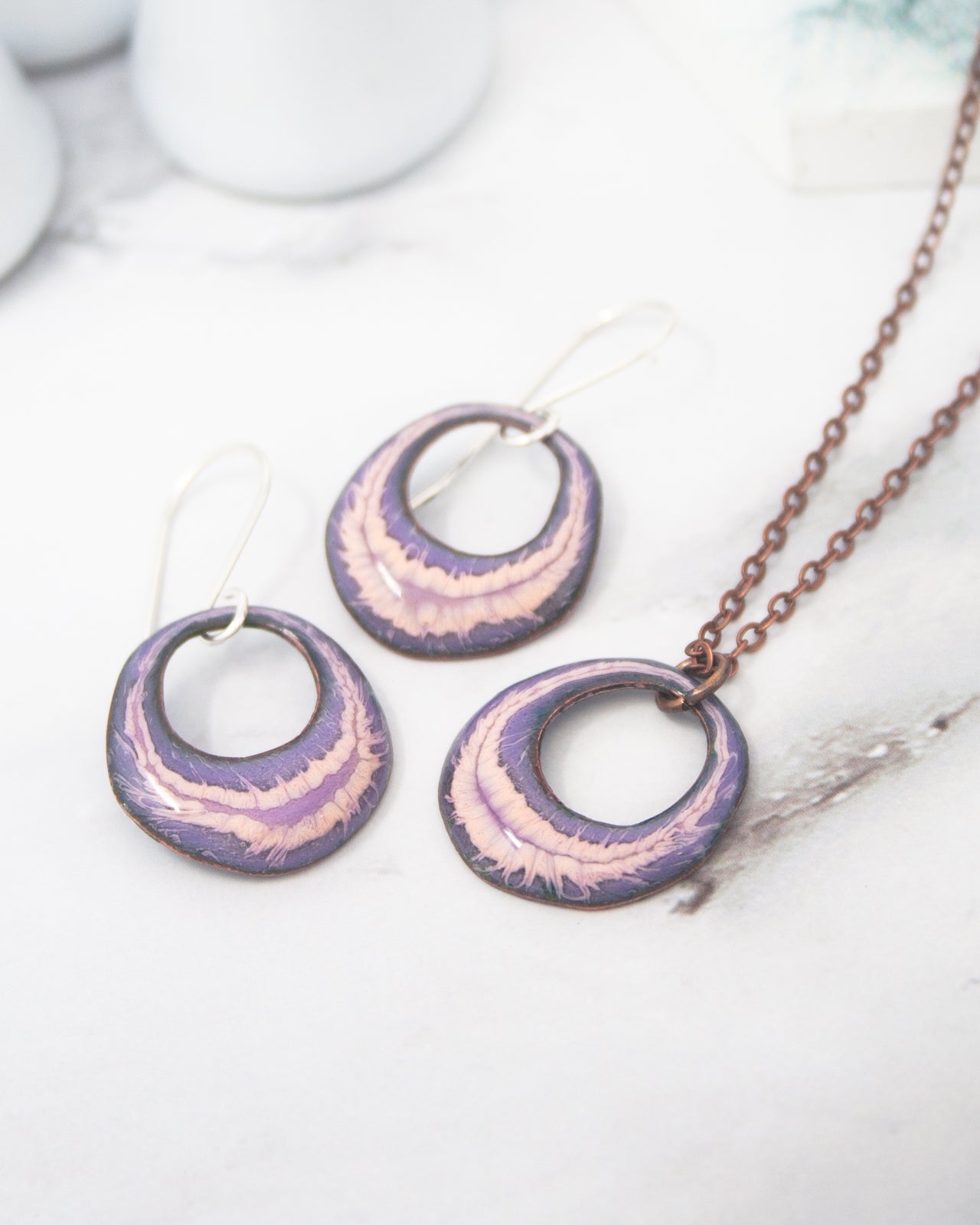 A necklace and earrings with purple and pink metallic designs are displayed on a white marble surface.