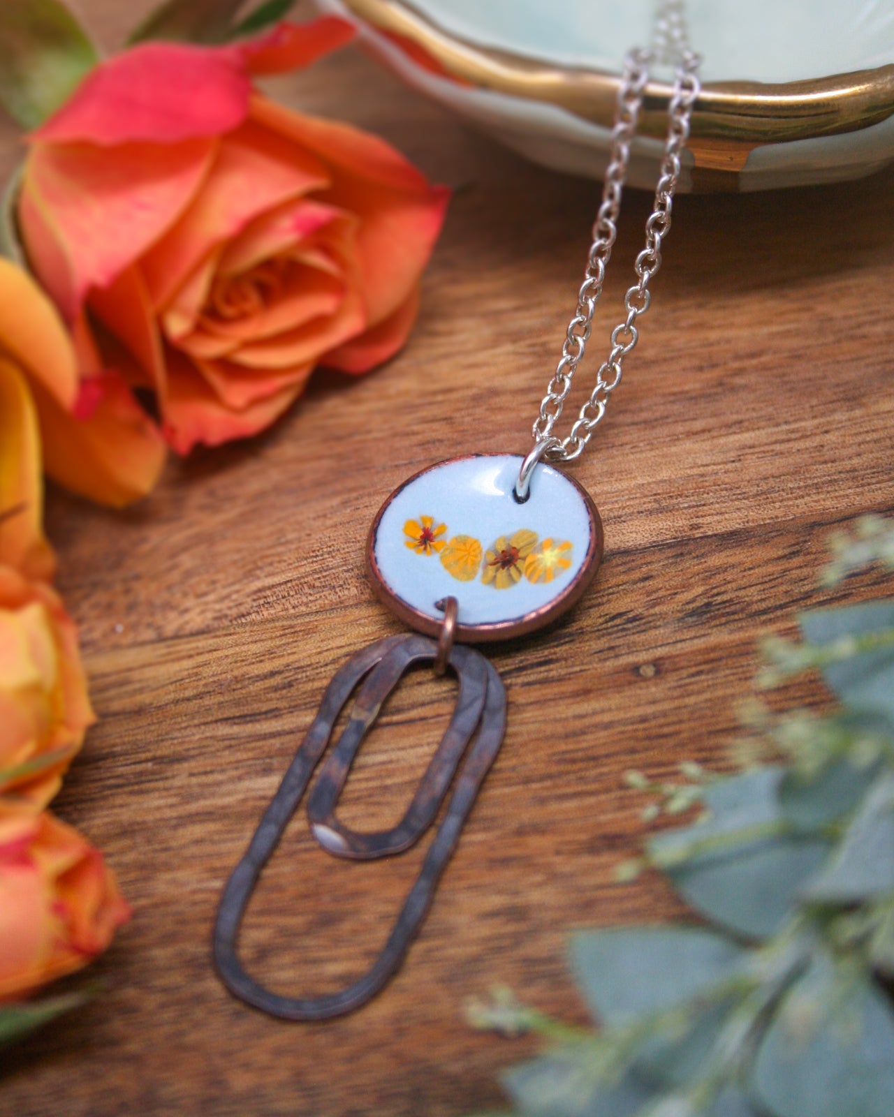 a necklace with a pendant that has a circular design with three yellow flowers on it, hanging from a chain. The necklace is placed on a wooden surface, with a bowl of flowers and a rose nearby.