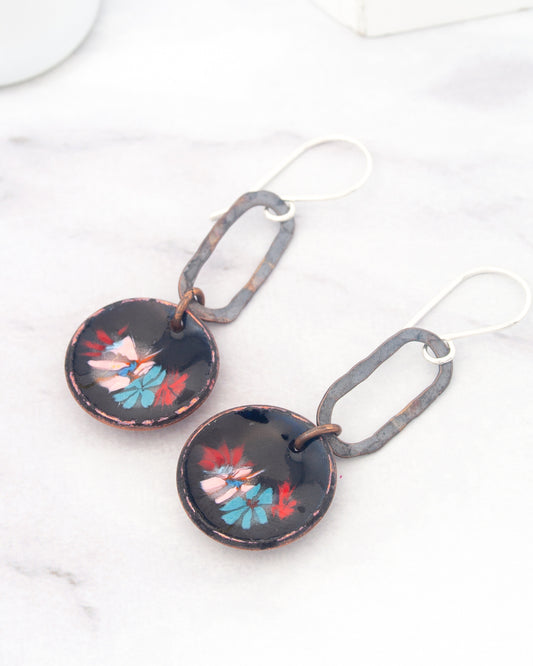A pair of earrings with a floral design on the circular pendants, hanging from a hook on a white marble surface.