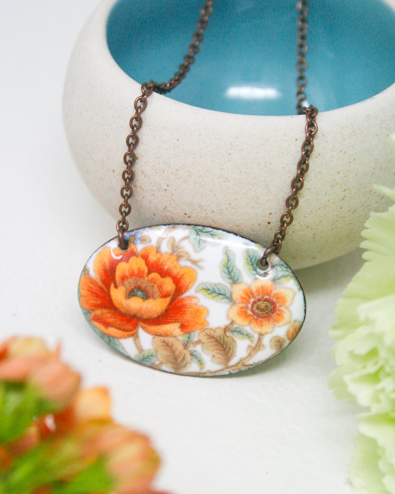 A necklace with a pendant featuring a floral design in orange and white colors, hanging from a chain. The pendant is displayed in front of a white bowl with a blue interior.