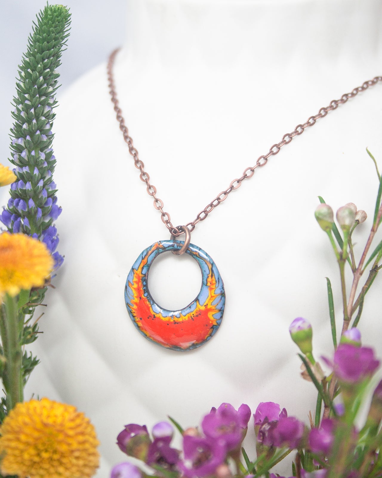 A necklace with a circular pendant featuring a vibrant design of blue and orange colors, surrounded by a variety of colorful flowers and foliage.