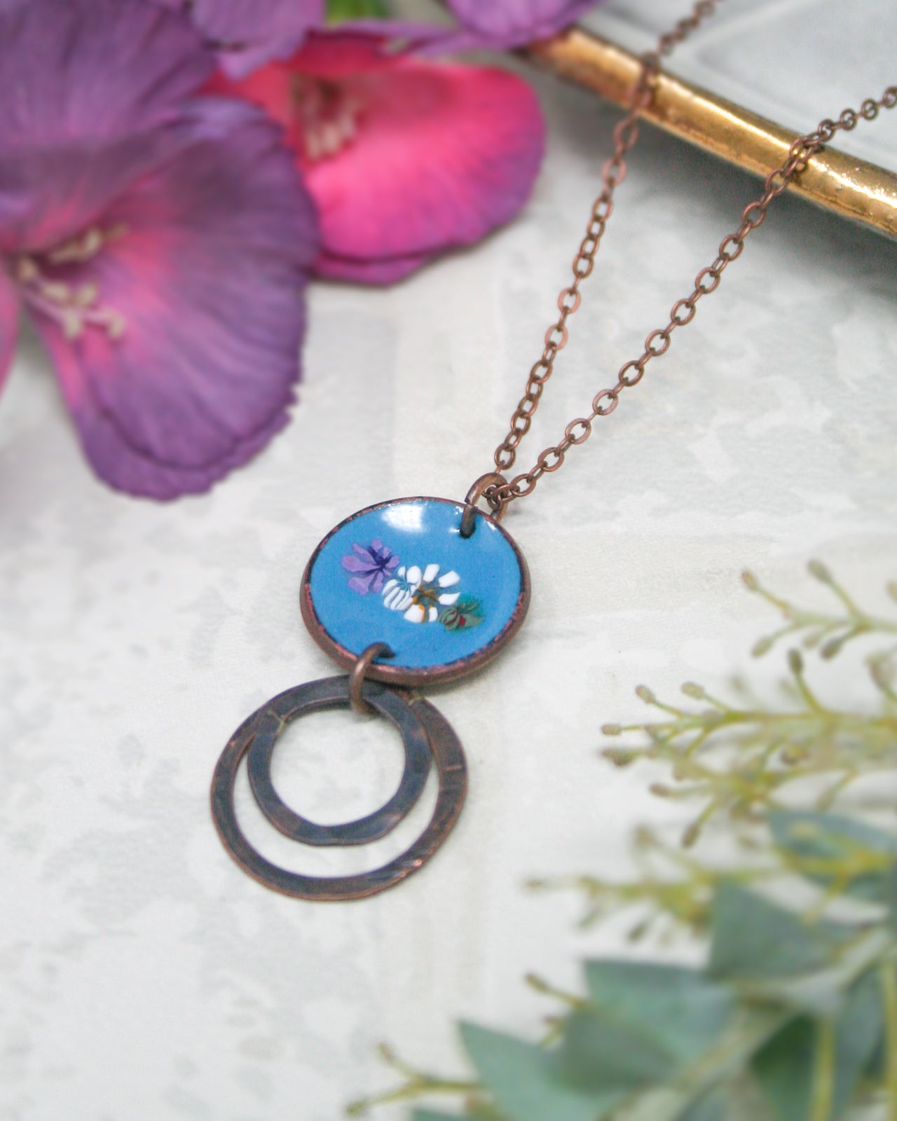 A blue pendant necklace with a circular design and two circular metal rings hangs from a chain, surrounded by purple and pink flowers and greenery.