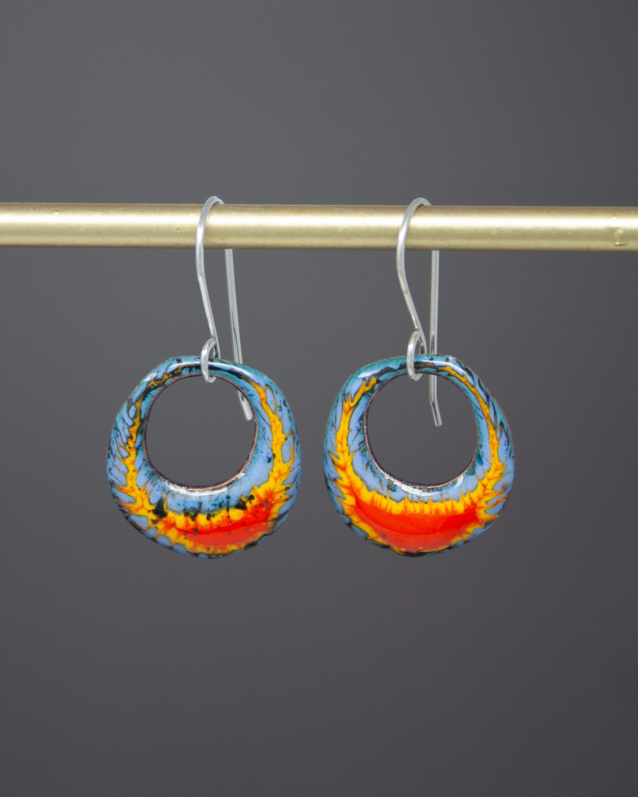 A pair of colorful earrings with blue, orange, and yellow hues hanging from a gold-colored metal bar.