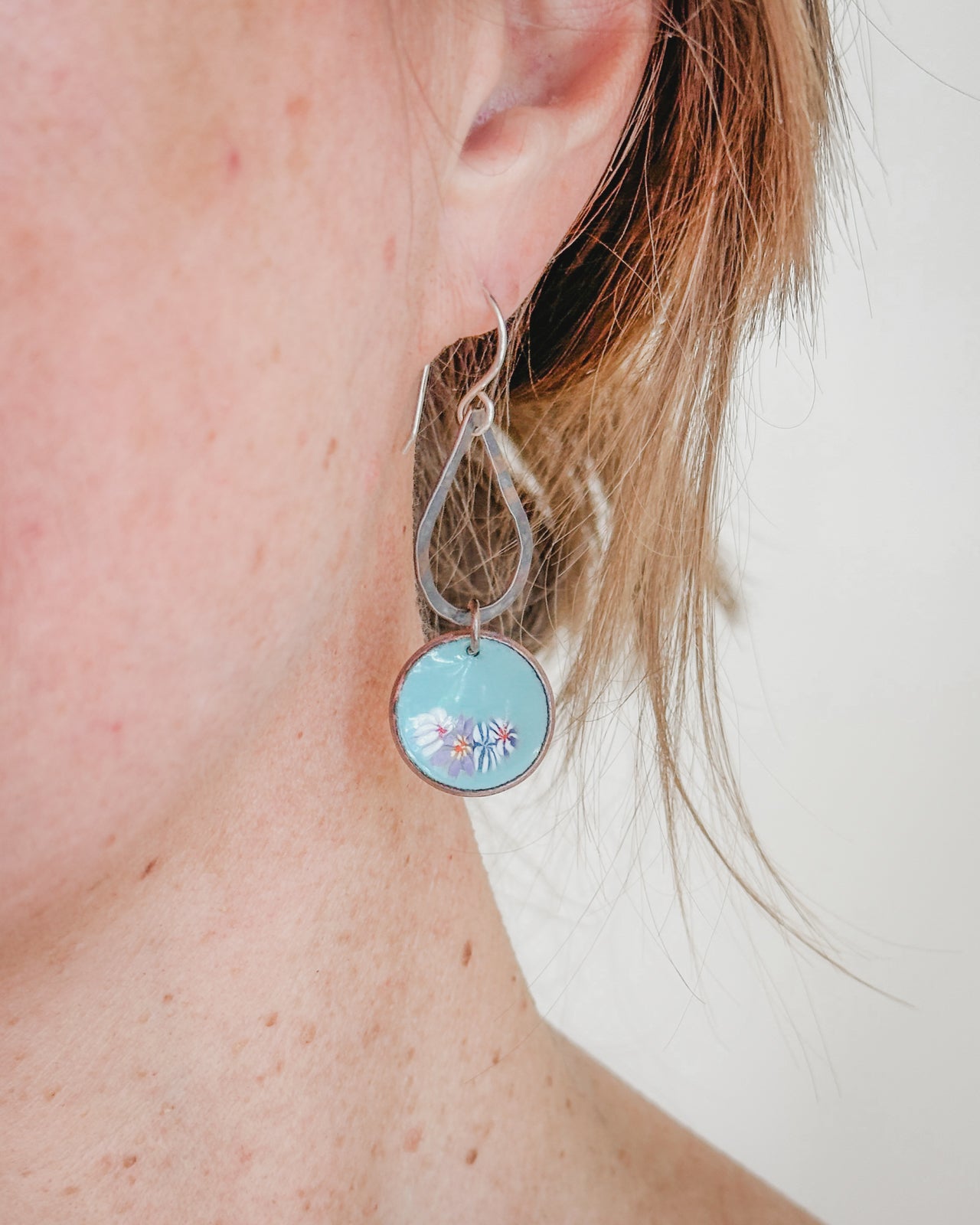 A close-up view of a person's ear wearing a pair of silver earrings with a blue circular charm featuring a floral design.