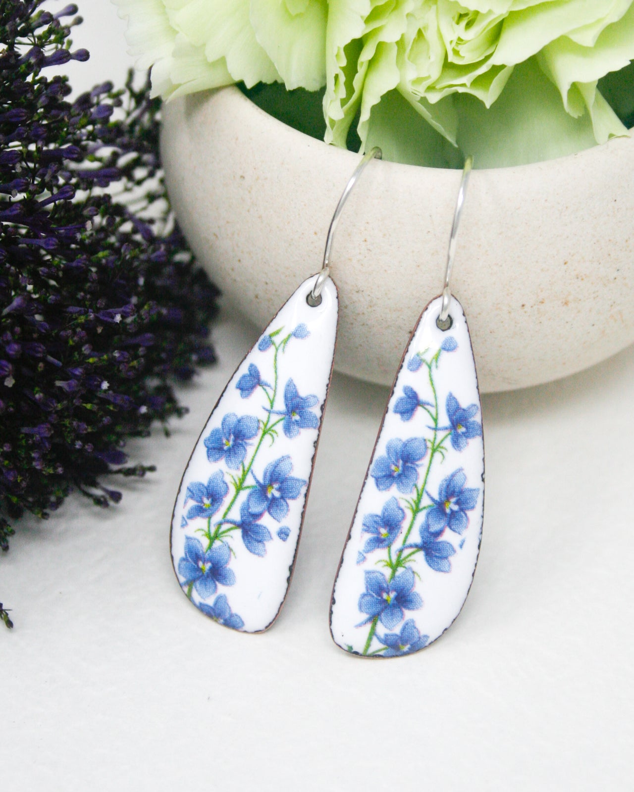 A pair of white earrings with blue flower designs, placed on a white surface next to a white bowl filled with green flowers.