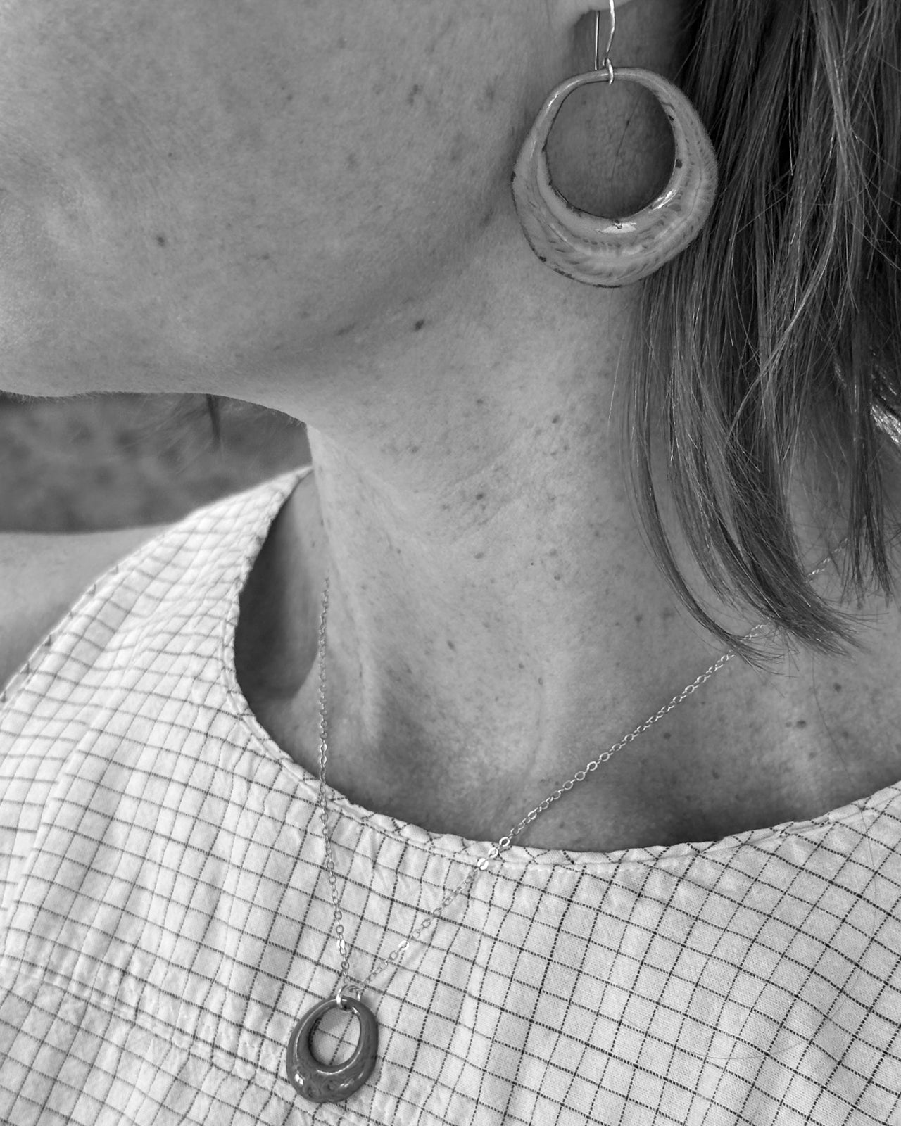 A close-up black and white image of a person's neck and chest area, featuring a necklace with a pendant and a large hoop earring.