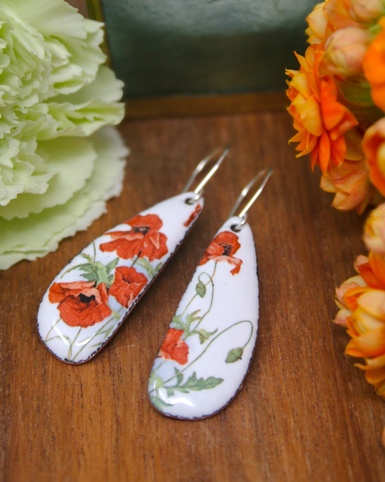 a pair of earrings with a floral design, placed on a wooden surface next to a bunch of orange flowers.