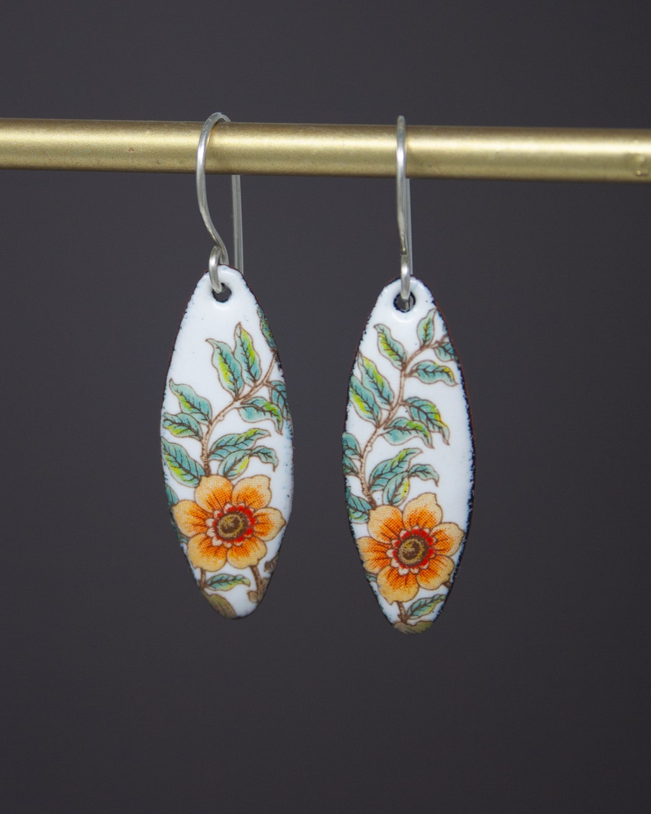 A pair of white earrings with a floral design, featuring orange and green flowers, hanging from a gold bar.